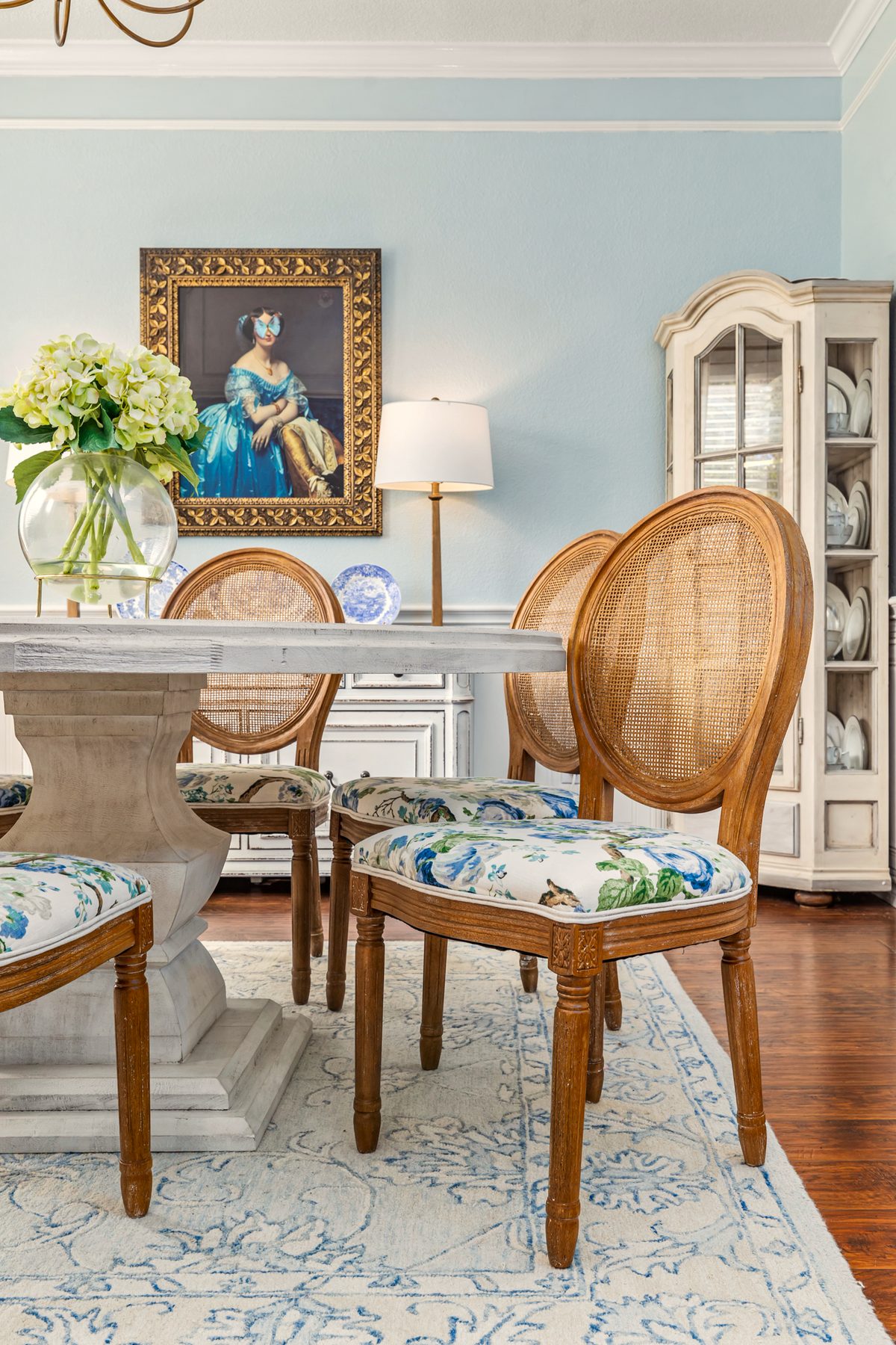 A blue dining room with cane chairs and a gilt-framed portrait, designed by Anna