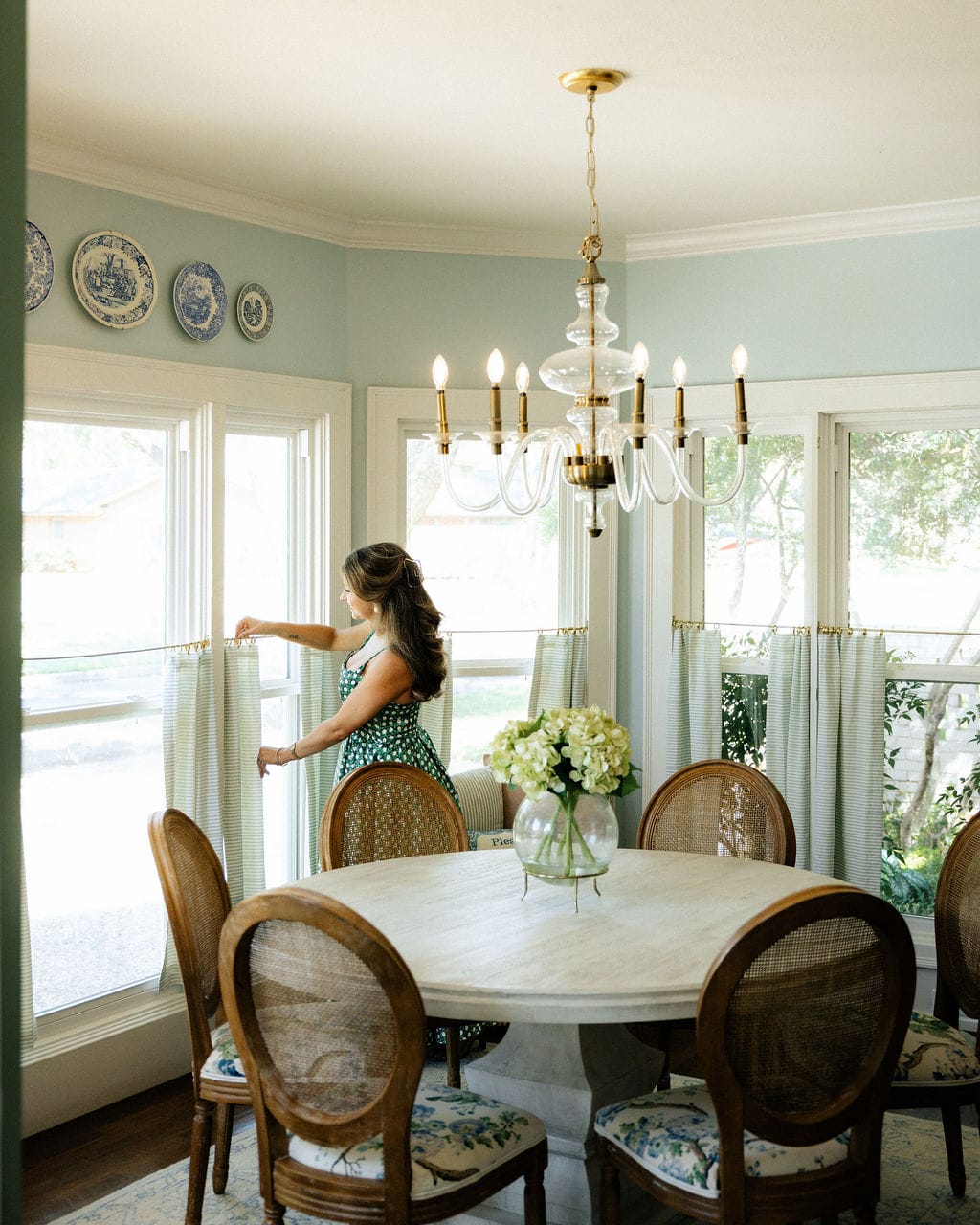 Anna Bouchelle adjusting cafe curtains in a blue dining room with cane chairs and a brass chandelier.