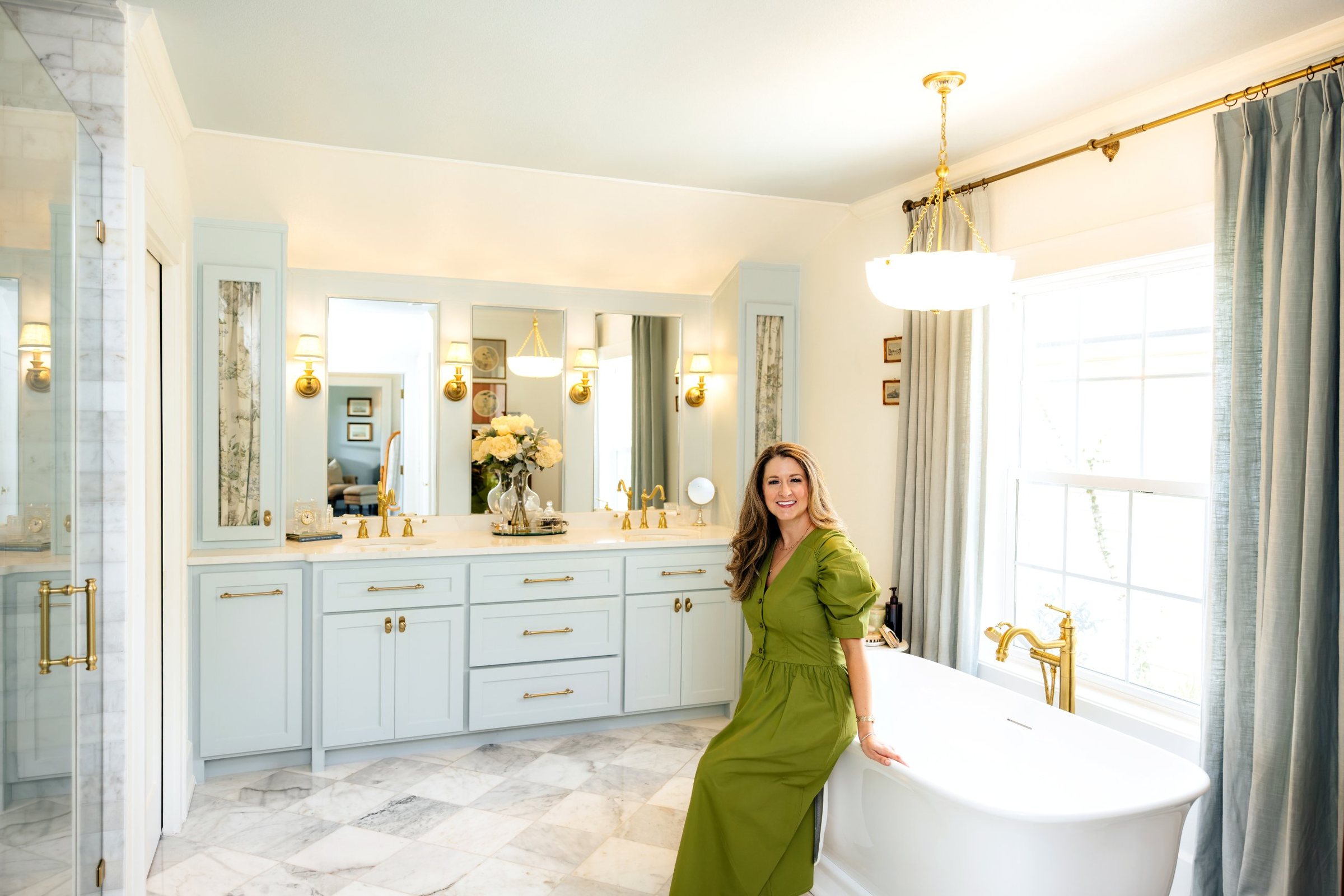 Anna Bouchelle in a green dress in a marble bathroom with a freestanding tub, pale blue cabinetry and brass fixtures.